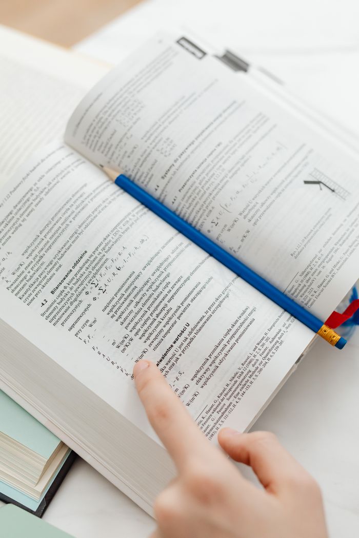 Close-up of a student's hand pointing at text in an open book with a pencil, emphasizing study.